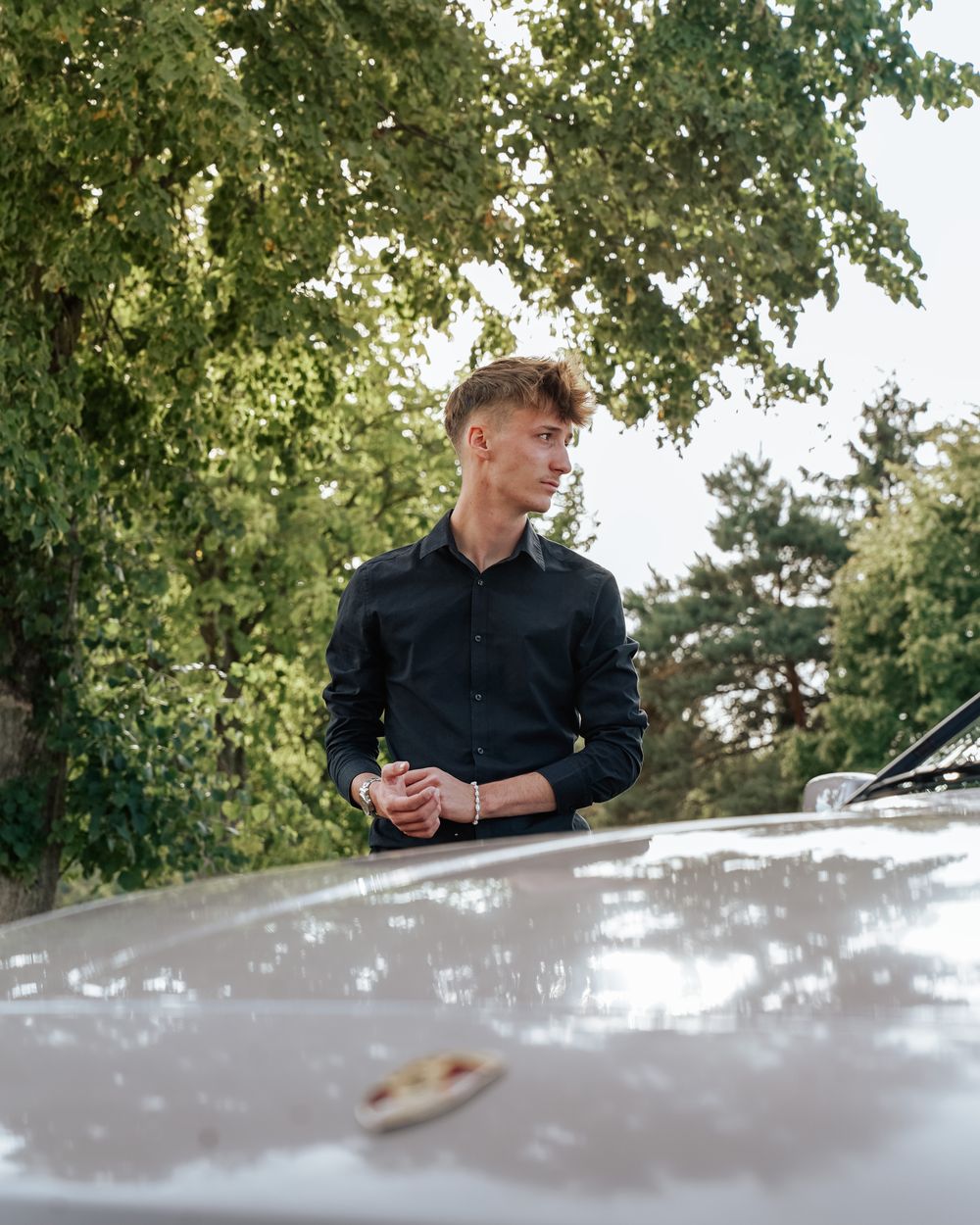 Mann im schwarzen Hemd vor einem Sportwagen unter Bäumen
